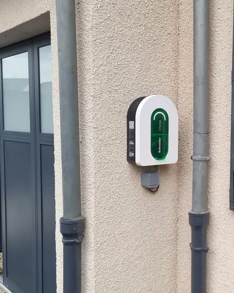 Borne de recharge Schneider Charge posée sur une façade crépie, entre deux descentes d’eau, à côté de la porte du garage, voyant vert allumé
