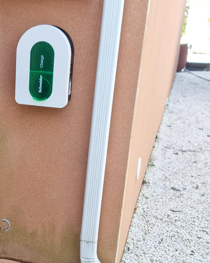 Borne de recharge murale Schneider Charge, blanche à vitre verte, posée dehors sur le mur enduit d’une maison, à côté d’une descente d’eau blanche ; à droite, l’angle de la maison et une allée de gravier