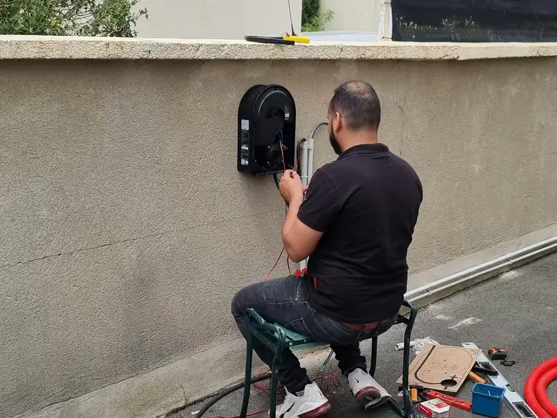 Technicien assis, de dos, qui raccorde une borne de recharge noire fixée sur un muret crépi coiffé d’une couvertine ; un tube gris longe le pied du muret, au bord d’une allée