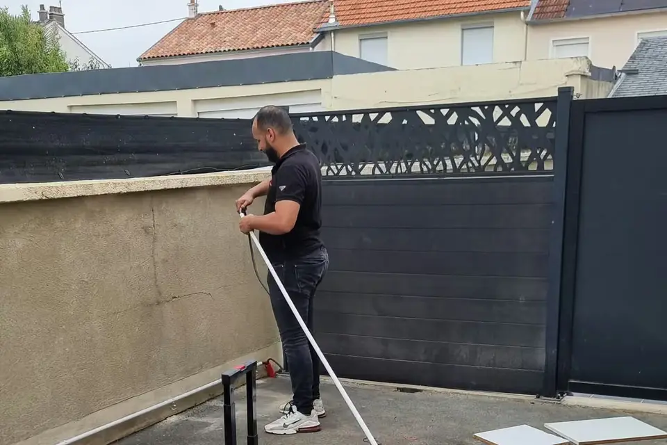 Technicien debout qui fait passer un câble dans un tube gris, le long d’un muret crépi ; clôture anthracite derrière