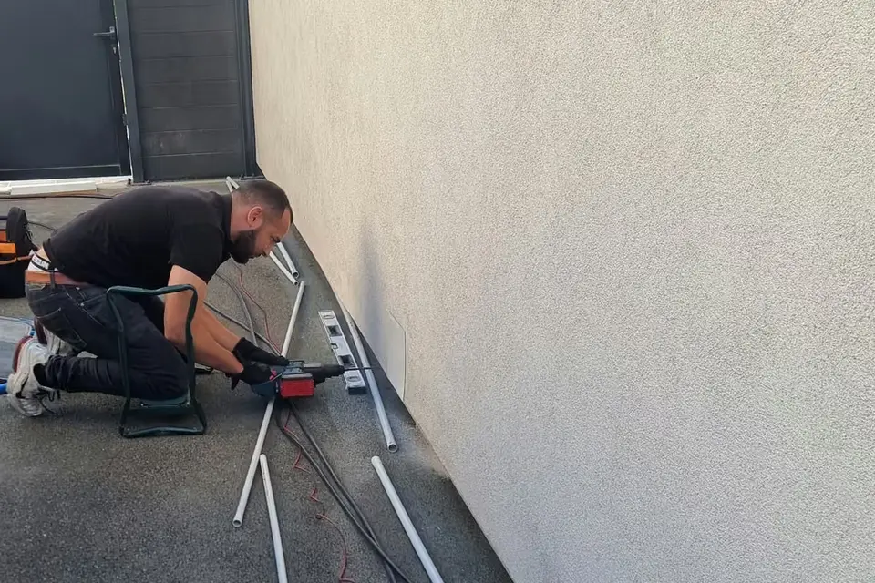 Technicien à genoux qui perce le bas d’une façade crépie ; tubes gris et câbles étalés au sol, clôture anthracite au fond