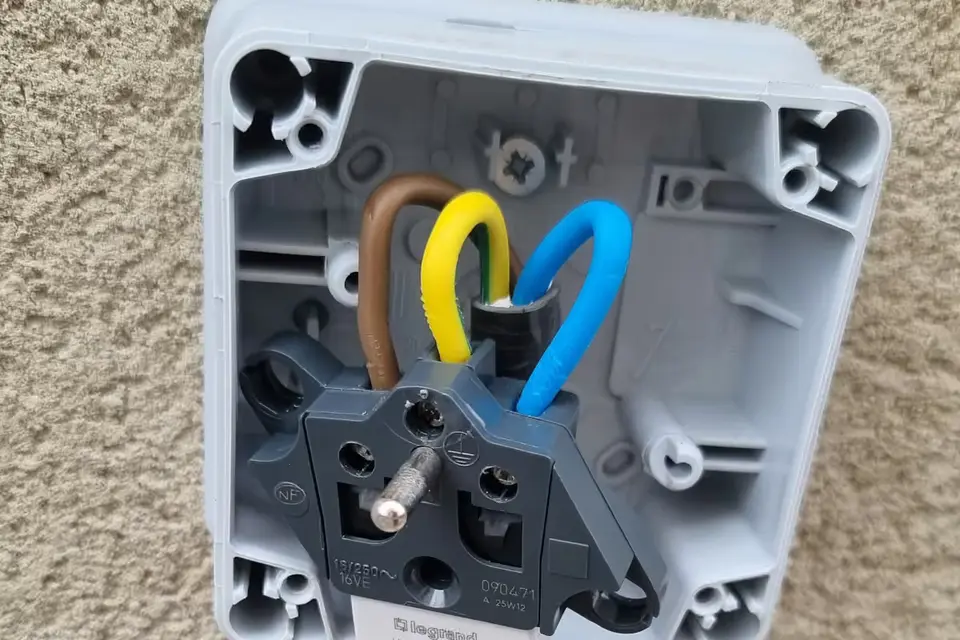 Prise renforcée étanche ouverte sur un mur crépi, fils marron, vert et jaune, et bleu raccordés