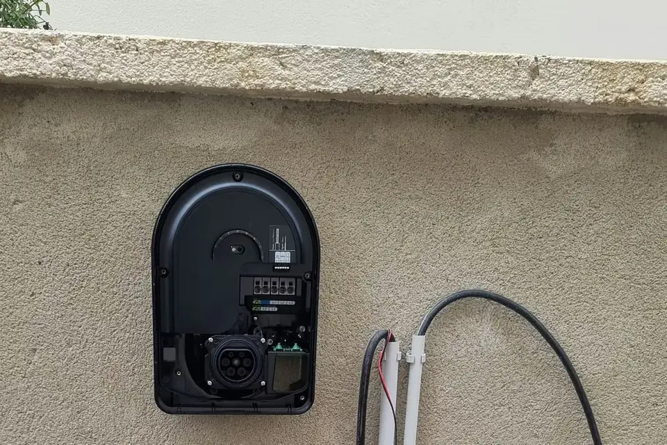 Socle noir d’une borne de recharge fixé sous la couvertine d’un muret crépi, capot retiré, prise Type 2 visible ; deux tubes gris et des câbles en attente à côté