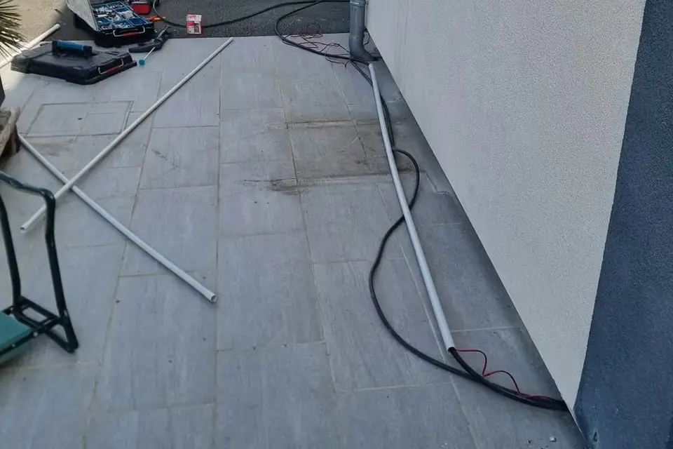 Tube gris et câble noir posés au pied d’une façade blanche, sur une terrasse dallée, jusqu’à une descente d’eau
