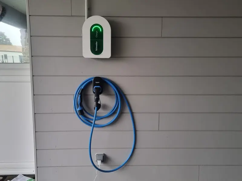 Borne de recharge murale blanche au voyant vert, posée dehors sur un bardage clair, câble bleu rangé sur son support