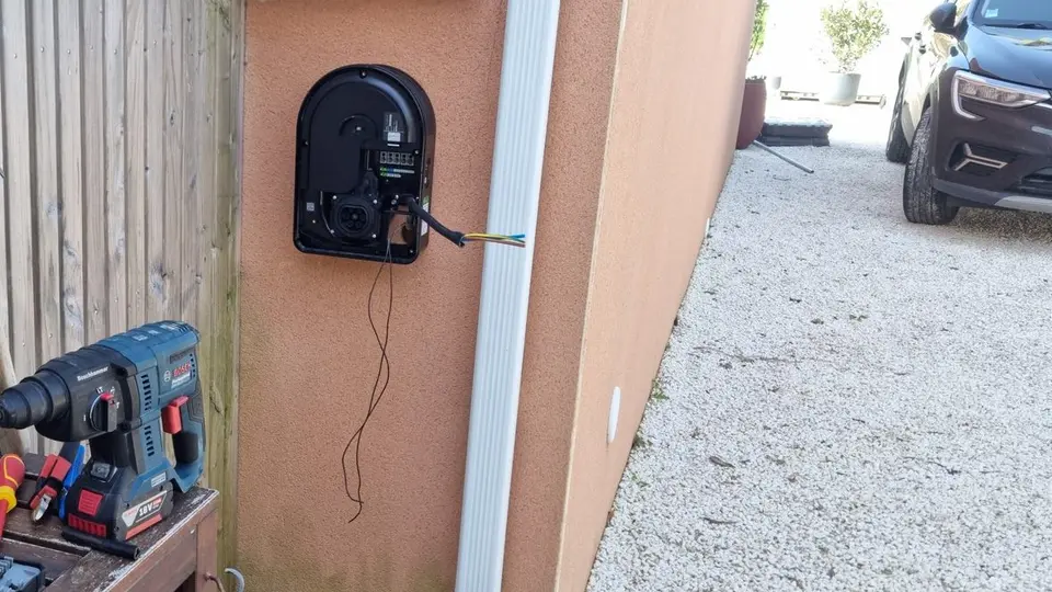 Borne de recharge ouverte, en cours de pose sur le mur d’une maison, perforateur posé sur une table de jardin à côté