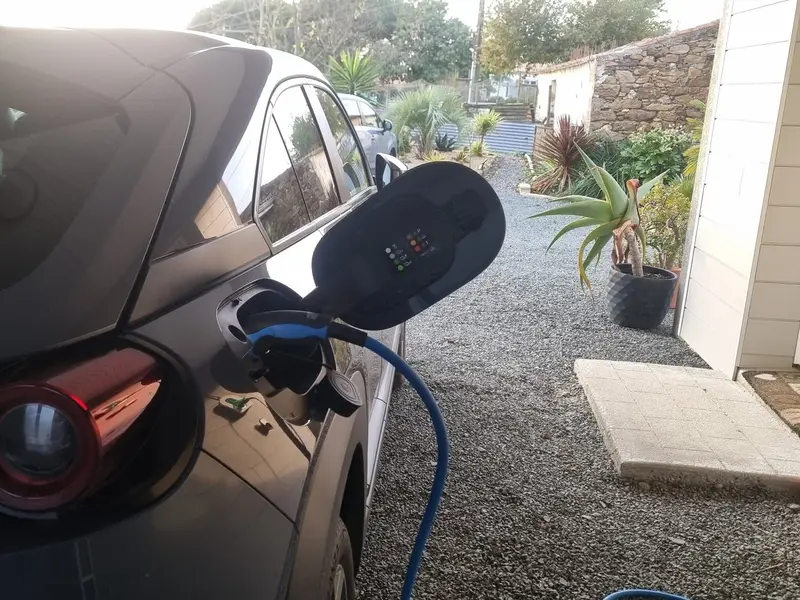 Arrière d’une voiture électrique grise en charge dans une cour gravillonnée, à côté d’une maison, câble bleu branché dans la trappe