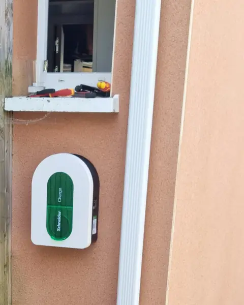 Borne Schneider Charge blanche à capot vert posée sur un mur crépi saumon, sous une fenêtre, près d’une descente d’eau blanche.