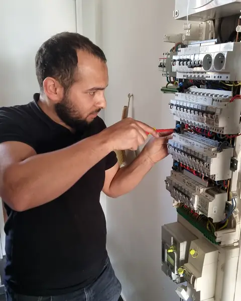 Un technicien d’Ave Green en polo noir intervient au tournevis isolé dans un tableau électrique ouvert, fixé à un mur blanc.