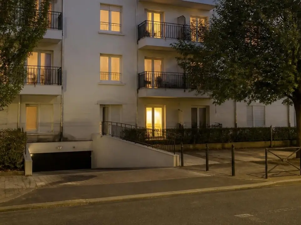 Façade d’une résidence au crépuscule, fenêtres allumées, rampe d’accès au parking souterrain
