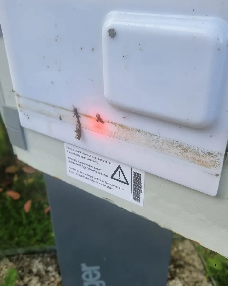 Capot blanc sali d’une borne de recharge sur pied Hager, voyant rouge allumé