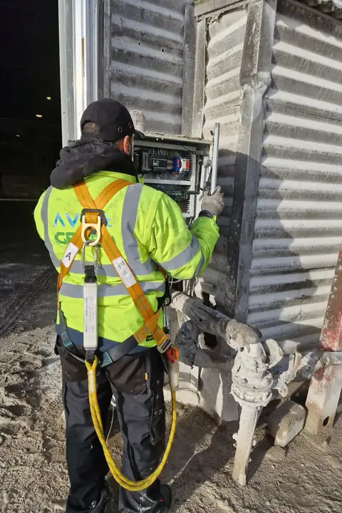 Électricien Ave Green de dos, en veste haute visibilité et harnais, devant une armoire électrique fixée sur un bardage