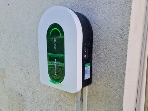 Borne de recharge murale blanche et noire, trappe verte translucide allumée, fixée sur un mur crépi ; son câble d’alimentation arrive par une goulotte blanche