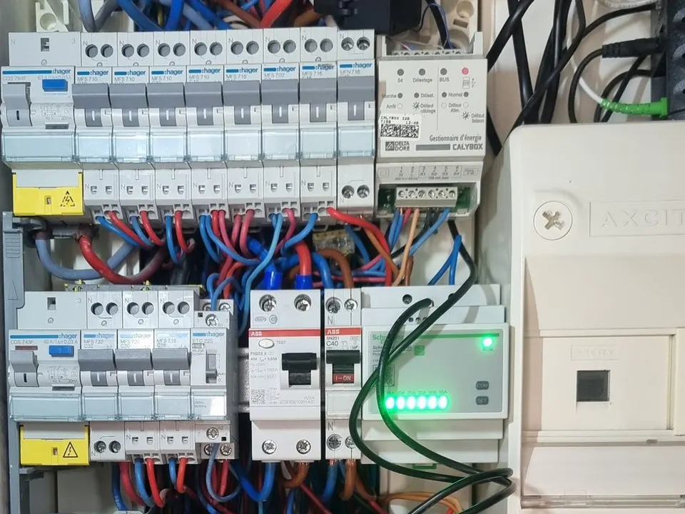 Tableau électrique ouvert : rangées de disjoncteurs, gestionnaire d’énergie en haut à droite et module à voyants verts allumés