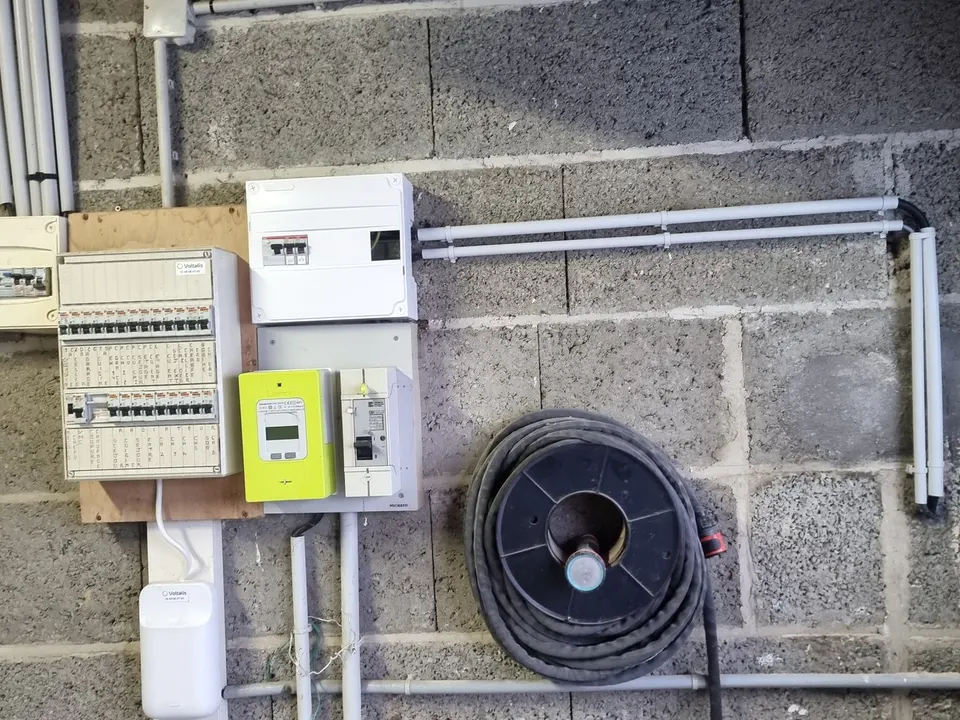 Compteur Linky vert, disjoncteur de branchement et tableau électrique sur un mur en parpaings, nouveau coffret blanc relié par deux conduits gris