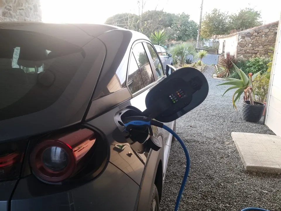 Voiture électrique gris foncé en charge dans une cour gravillonnée en fin d’après-midi, câble bleu branché dans sa trappe de recharge
