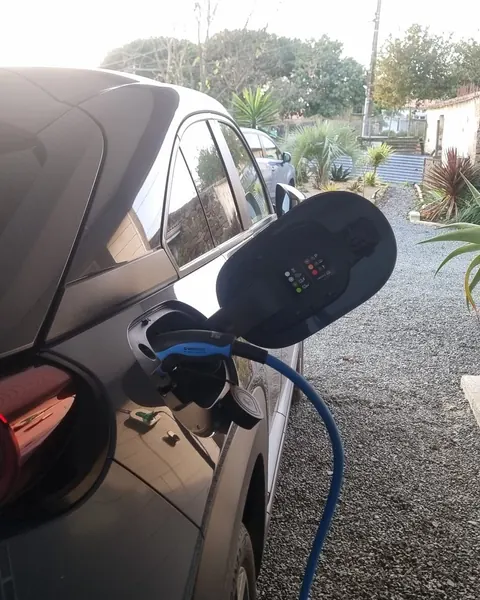 Voiture électrique en charge dans une cour gravillonnée en fin d’après-midi, câble bleu branché dans sa trappe de recharge