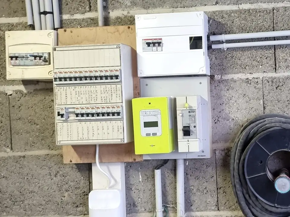 Ancien tableau électrique étiqueté à la main, compteur Linky et disjoncteur de branchement sur un mur en parpaings ; au-dessus, un coffret blanc ajouté, relié par des conduits gris