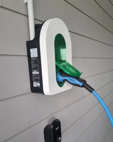 Borne murale blanche à prise Type 2 sur un bardage gris, clapet vert ouvert, fiche d’un câble de recharge bleu branchée dans la prise