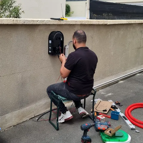 Le technicien, de dos, raccorde une borne murale sur un muret crépi, ses outils posés au sol