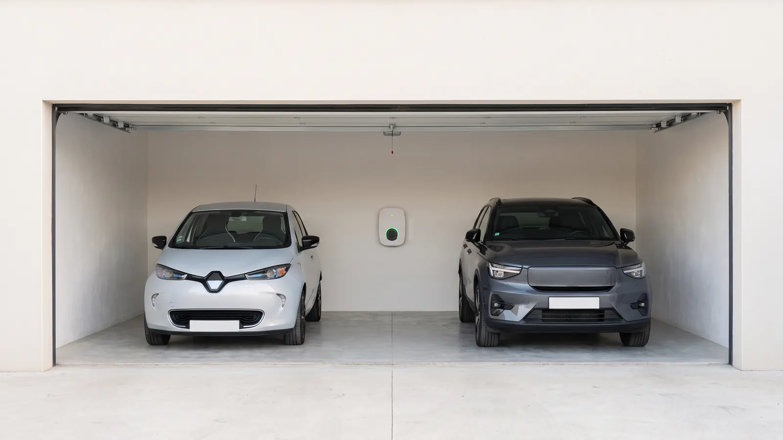 Garage ouvert d’une maison : une citadine électrique blanche et un SUV électrique gris garés de face, de part et d’autre d’une borne de recharge murale blanche