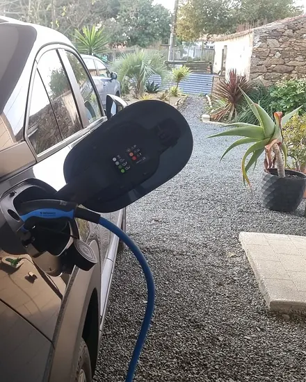 Flanc d’une voiture gris foncé en charge : trappe ouverte, prise branchée au câble bleu, cour gravillonnée