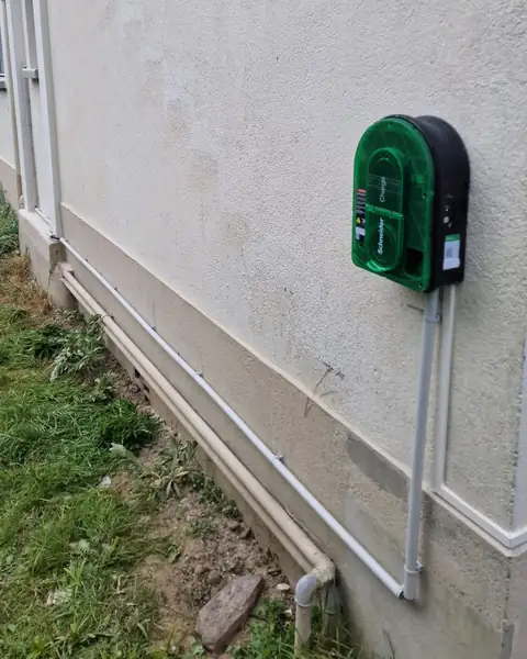 Borne Schneider Charge verte fixée sur une façade crépie claire&nbsp;; deux tubes gris longent le soubassement jusqu’à la borne