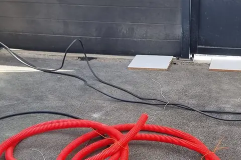 Gaine rouge enroulée au sol et câbles noirs posés sur l’enrobé, avant le passage sous l’allée