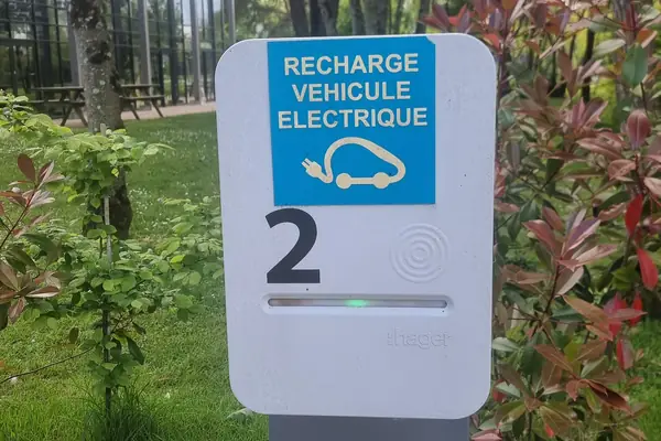 Borne de recharge sur pied numérotée 2, autocollant bleu « Recharge véhicule électrique », voyant vert allumé, entre des massifs de photinias rouges et une pelouse