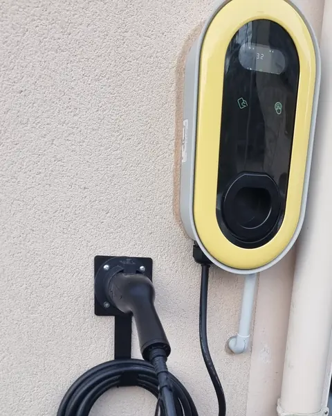 Borne de recharge murale à cadre jaune et écran noir posée dehors sur le mur enduit d’une maison, près d’une descente d’eau ; le pistolet est rangé sur son support et le câble noir est enroulé dessous