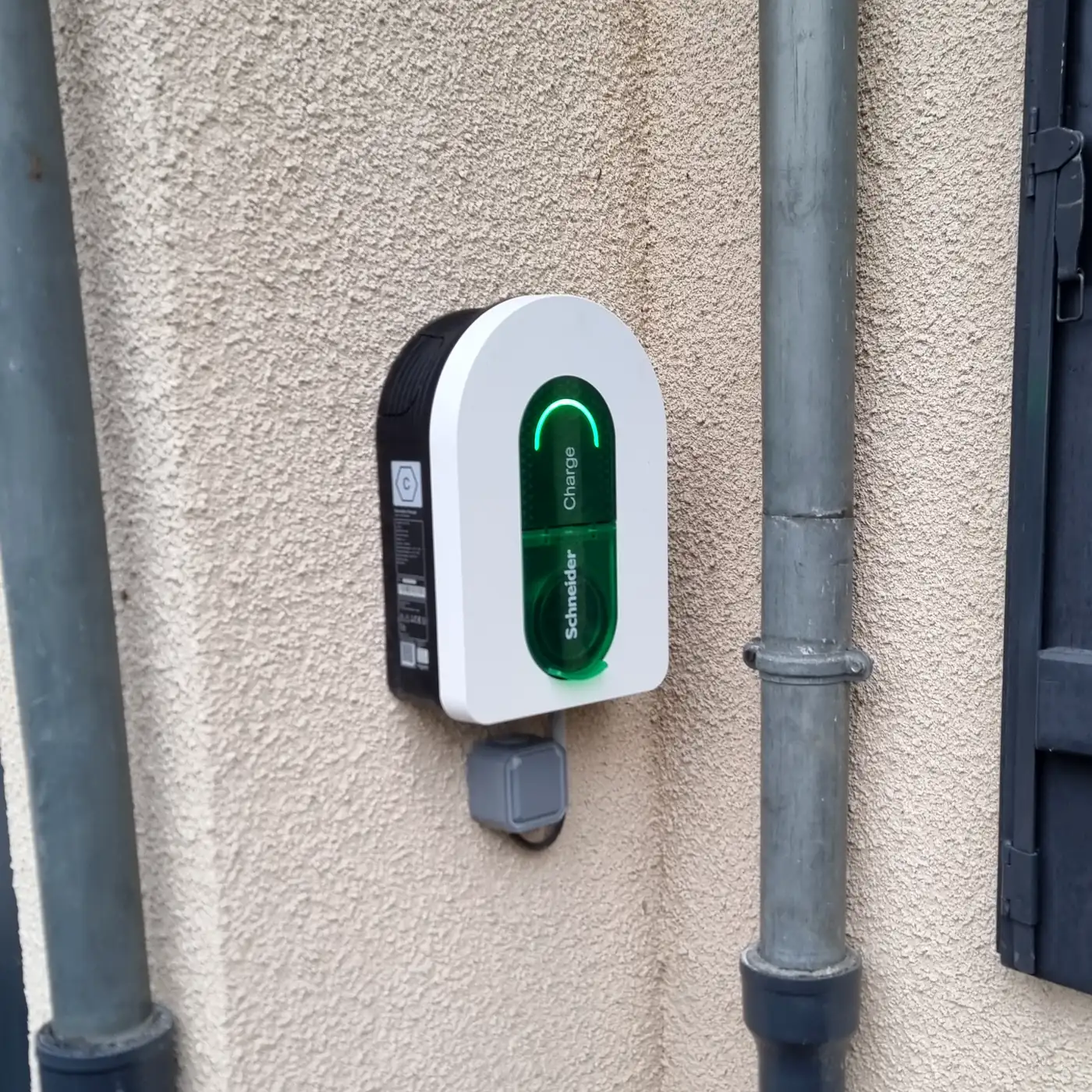 Borne de recharge Schneider Charge blanche, voyant vert allumé, fixée sur la façade crépie beige d’une maison, à côté d’une descente d’eau