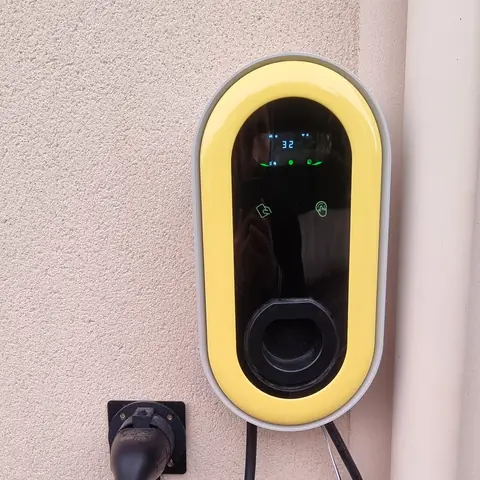 Borne de recharge murale ovale à cadre jaune, écran allumé affichant 32, fixée sur la façade enduite d’une maison, pistolet rangé sur son support, à côté d’une descente d’eau