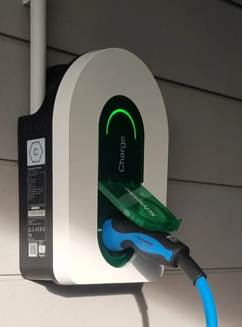 Borne de recharge murale Schneider Charge posée dehors sur un bardage clair, au soleil : voyant vert allumé, clapet vert ouvert, prise branchée sur un câble bleu