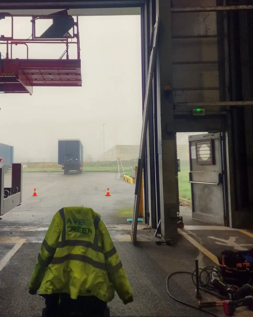 Veste haute visibilité Ave Green posée sur une caisse à l’entrée d’un entrepôt, parking dans la brume